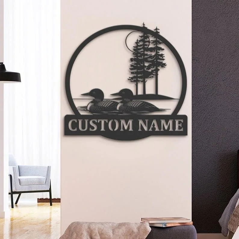 Black metal wall sign with two loons on water, pine trees and moon above, and placeholder text 'CUSTOM NAME', mounted on a living room wall