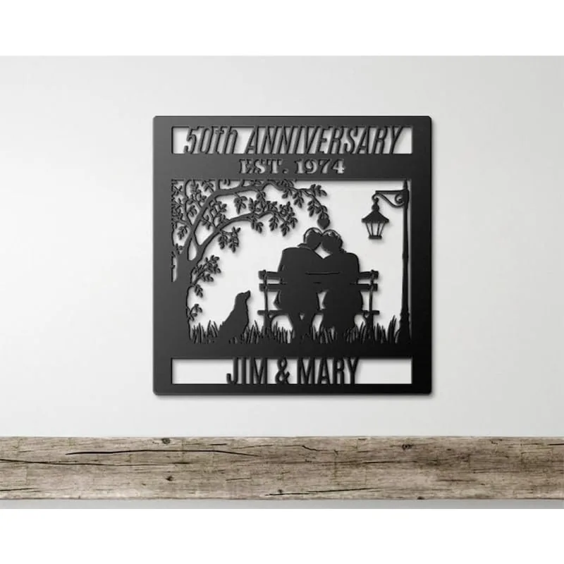 Black metal wall plaque reading "50th Anniversary Est. 1974" with silhouettes of a couple on a bench, a dog, tree, lamp, and names "Jim & Mary" hung on a white wall above a wooden shelf